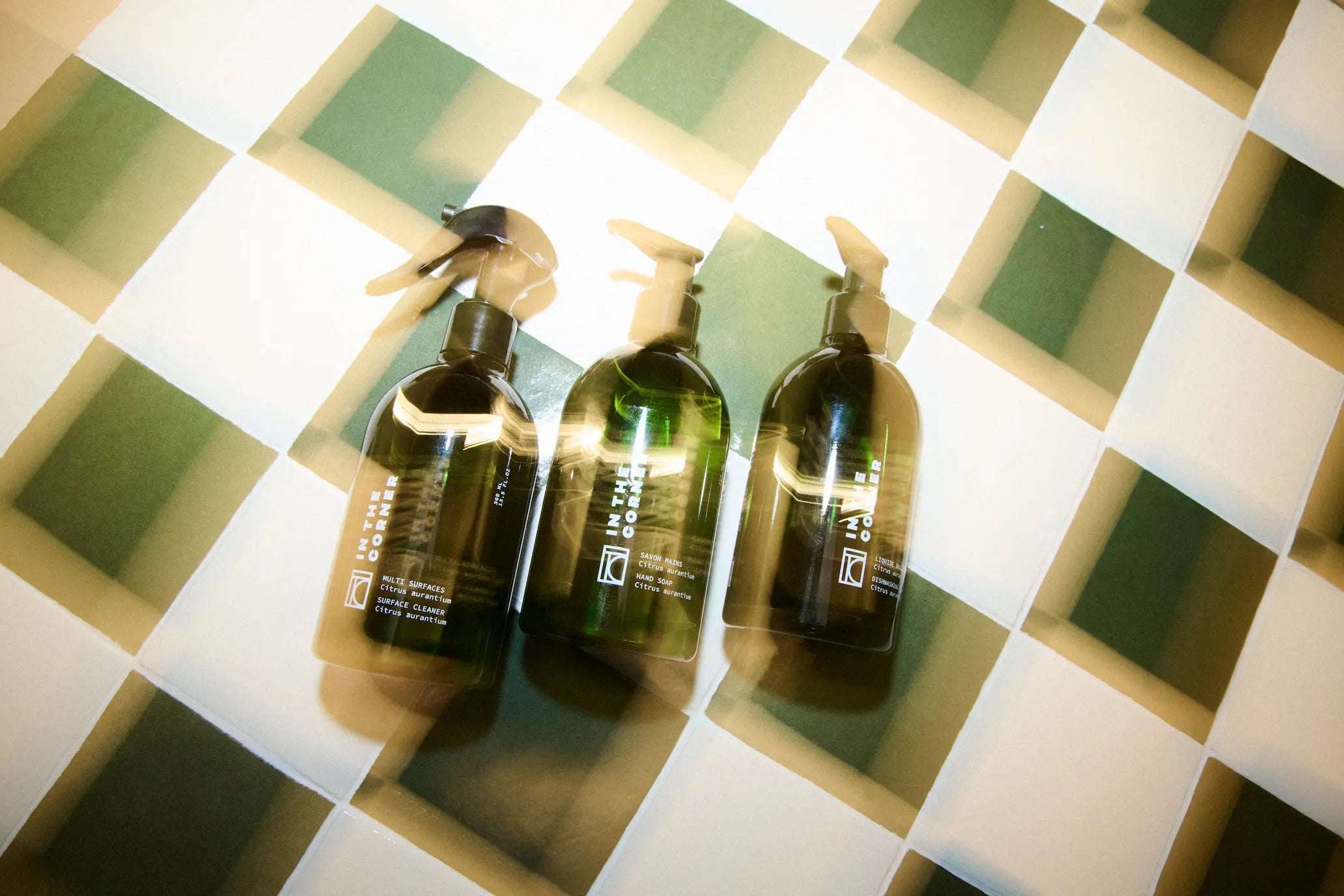 Trio de produits In the Corner posé sur une table à carreaux, composé du nettoyant multisurfaces, savon corps et main, liquide vaisselle, dans de jolies bouteilles vertes au design épuré