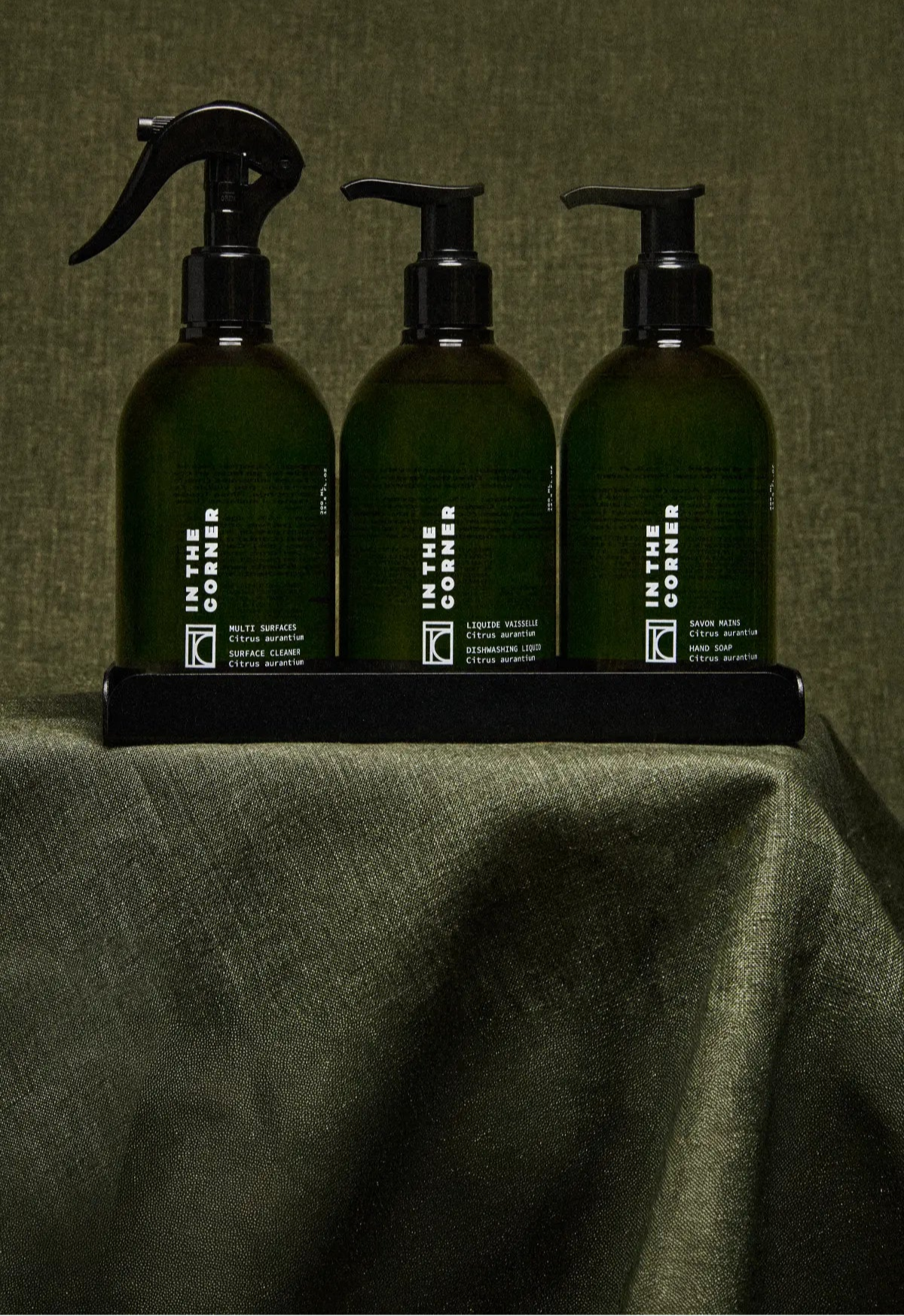 Trio de produits In the Corner posé sur une table, avec un fond et une nappe verte, composé du nettoyant multisurfaces, savon corps et main, liquide vaisselle, dans de jolies bouteilles vertes au design épuré