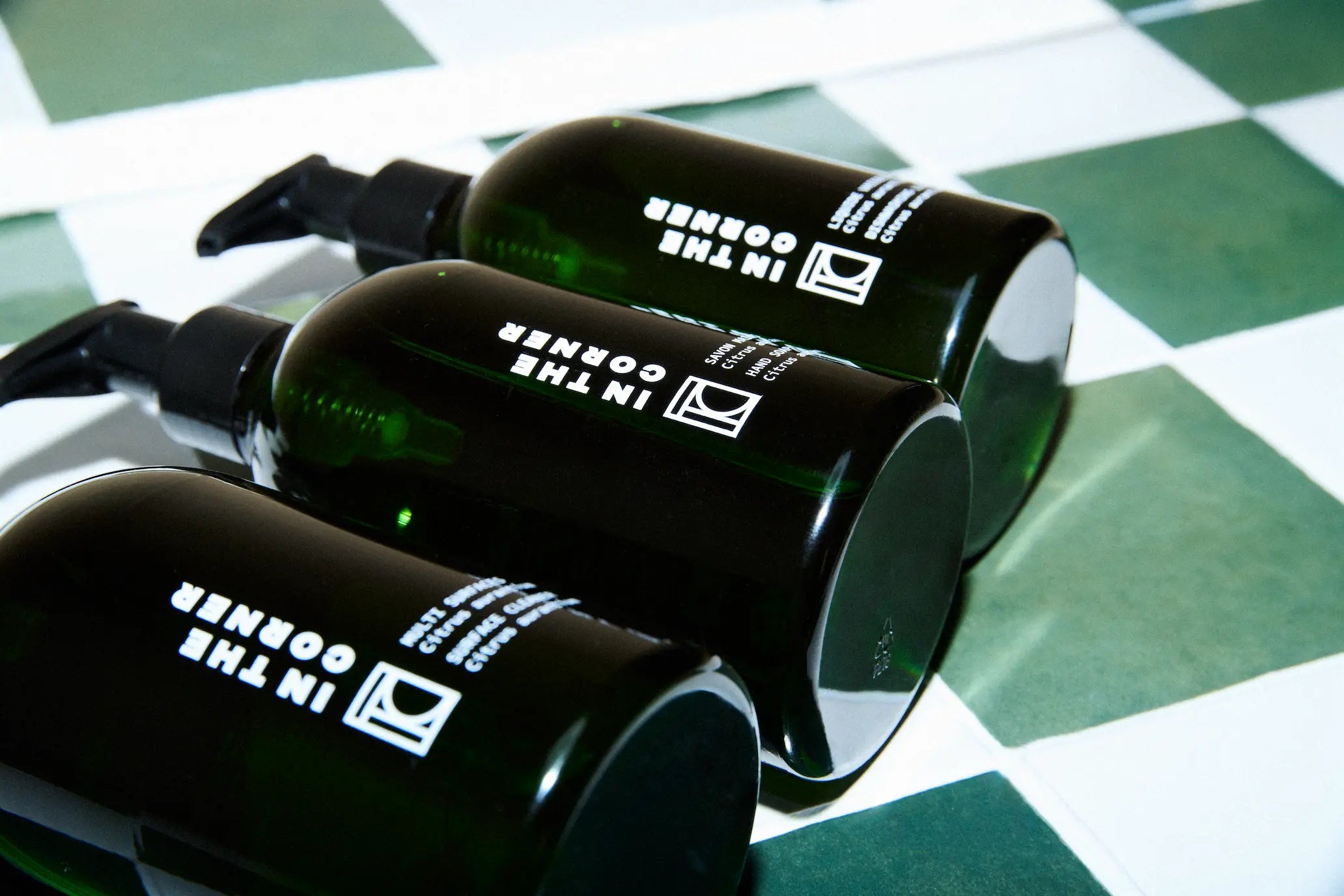 Trio de produits composé du nettoyant multisurfaces, savon corps et main, liquide vaisselle, dans de jolies bouteilles vertes au design épuré et élégant, couchés sur un fond en carrelage vert et blanc