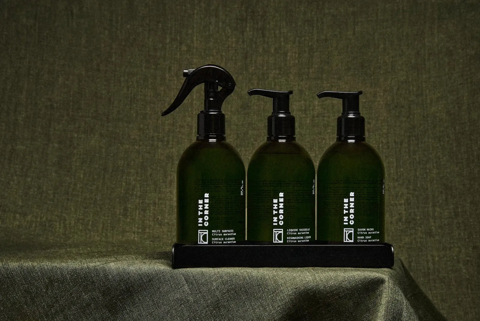 Trio de produits In the Corner posé sur une table, avec un fond et une nappe verte, composé du nettoyant multisurfaces, savon corps et main, liquide vaisselle, dans de jolies bouteilles vertes au design épuré